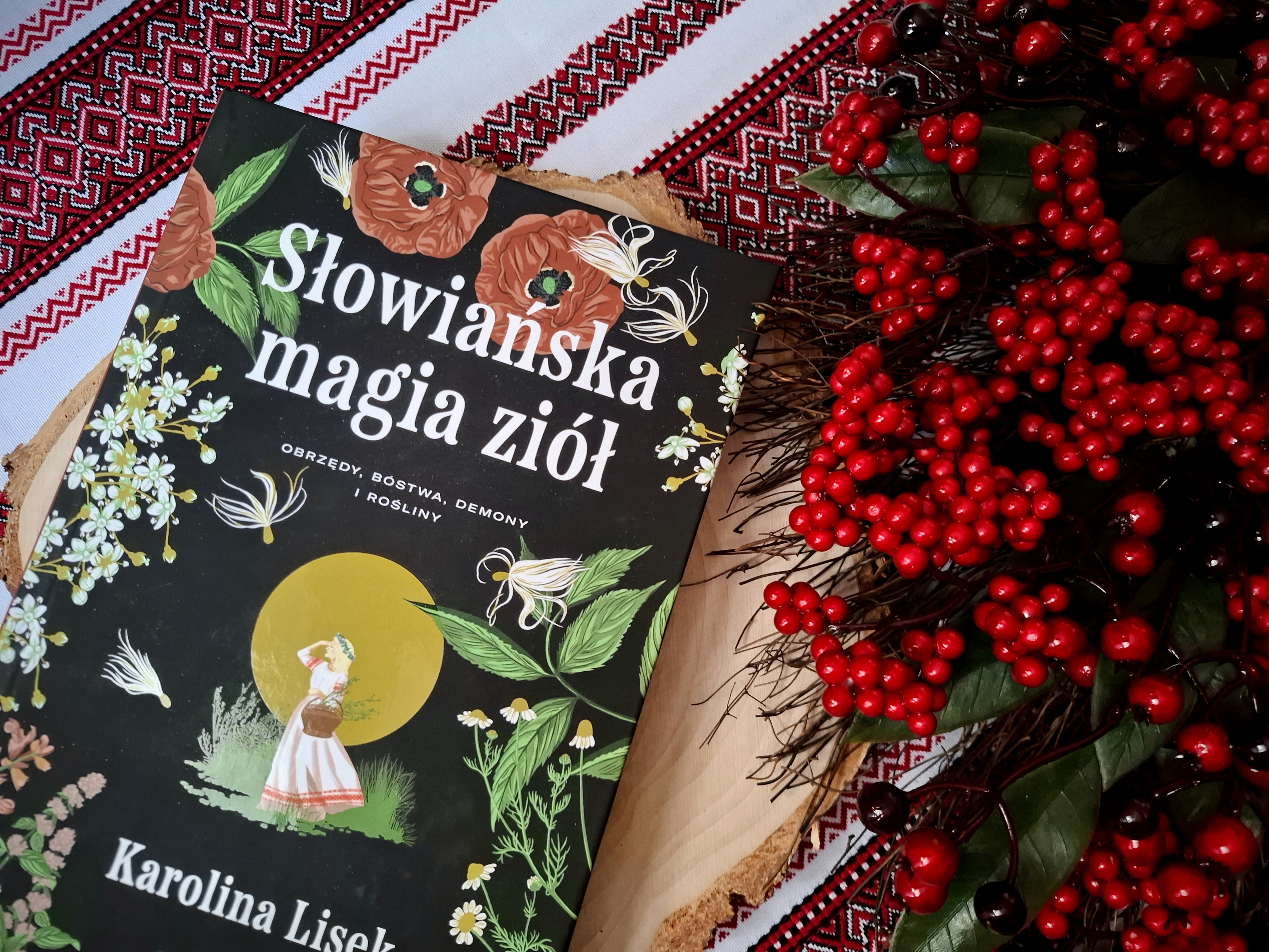 Zbliżenie na okładkę książki "Słowiańska magia ziół" - okładka ma czarne tło, na brzegach znajdują się motywy roślinne, pośrodku żółty księżyc i postać słowiańskiej dziewczyny, a także zapisany białymi literami tytuł książki i nazwisko autorki. Książka leży na plastrze brzozy, obok leży wianek z czerwonego głogu. Wianek i książka leżą na białym obrusie w czerwone hafty